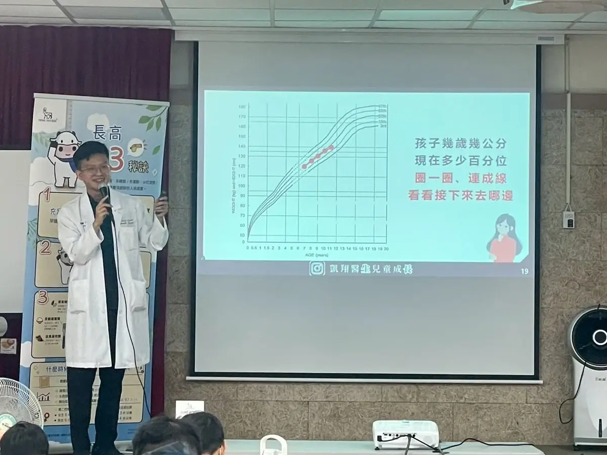 張凱翔醫師拿著麥克風，在投影幕旁示範如何在生長曲線圖上畫出孩子的身高