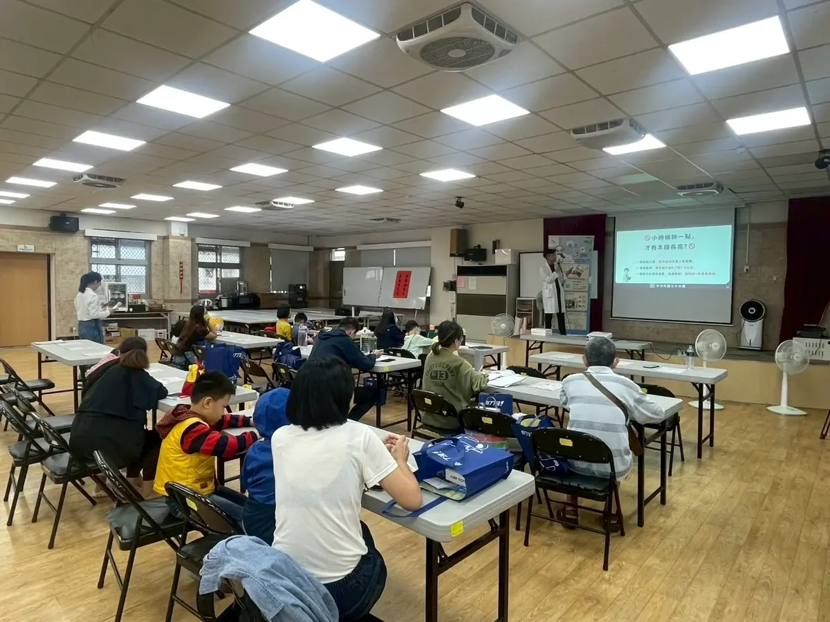 社區活動中心的講座現場，家長與孩子坐在長桌前聽講，張凱翔醫師在前方的投影幕旁說明