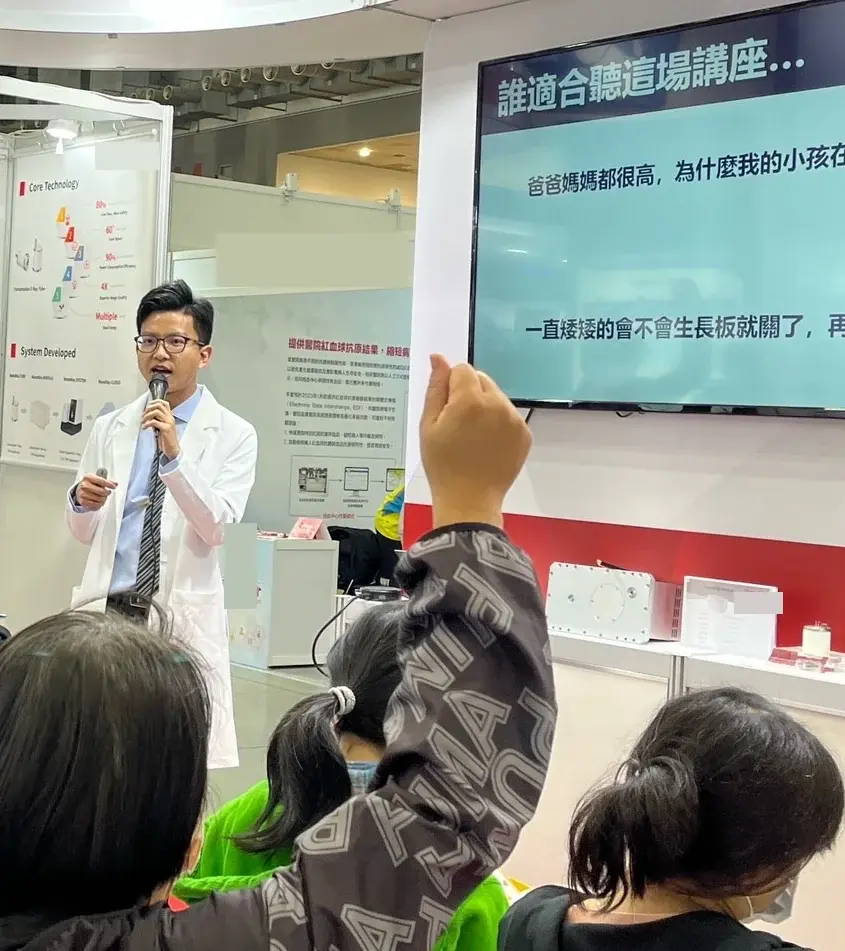 張凱翔醫師在展場講台旁拿著麥克風演講，台下家長舉手回應